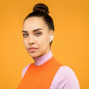 Portrait of a trendy woman wearing earbuds against a vibrant orange background, showcasing modern fashion.