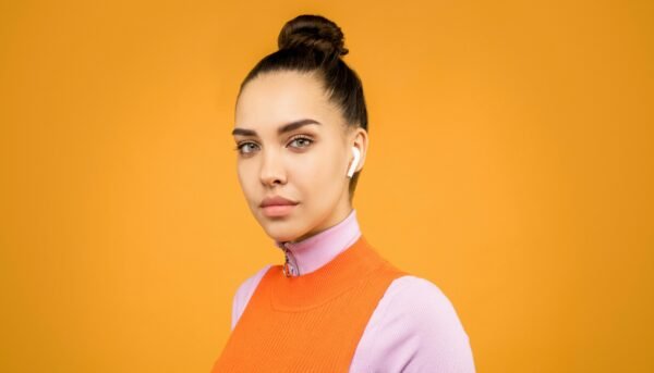 Portrait of a trendy woman wearing earbuds against a vibrant orange background, showcasing modern fashion.