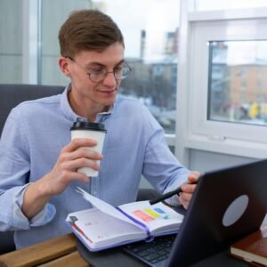 Business professional sipping coffee while working on a laptop with a planner at an office.