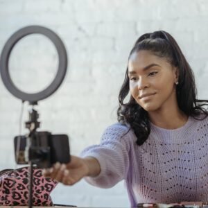 Glad African American female blogger with long dark hair using mobile phone and ring lamp while having video conversation online